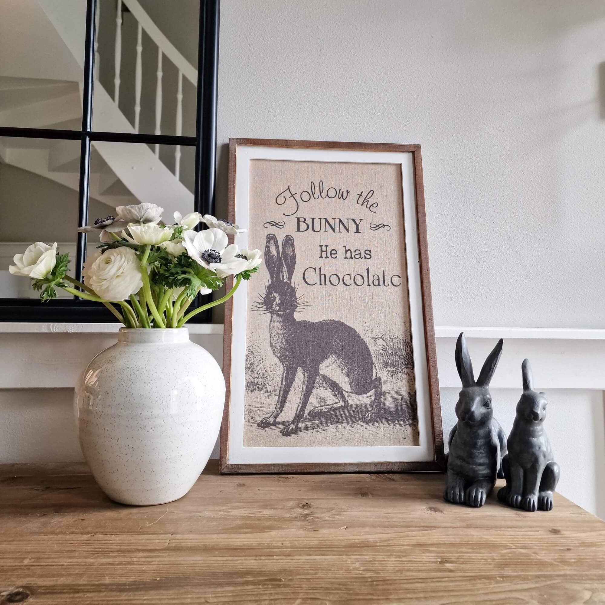 Dekoratives Bild mit Hasenmotiv und Schrift, weisse Blumen in Keramikvase, zwei Hasenfiguren auf Holztisch