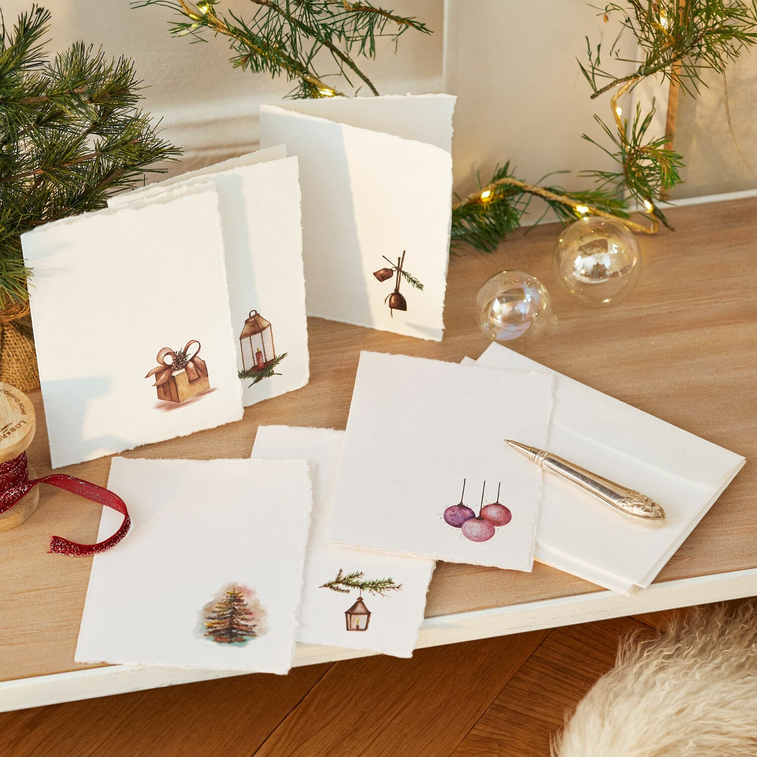 Grusskarten 6er Set Aeloria Set aus weissen Weihnachtskarten mit Tannen- und Geschenk-Motiven auf Holztisch mit LED-Lichterkette