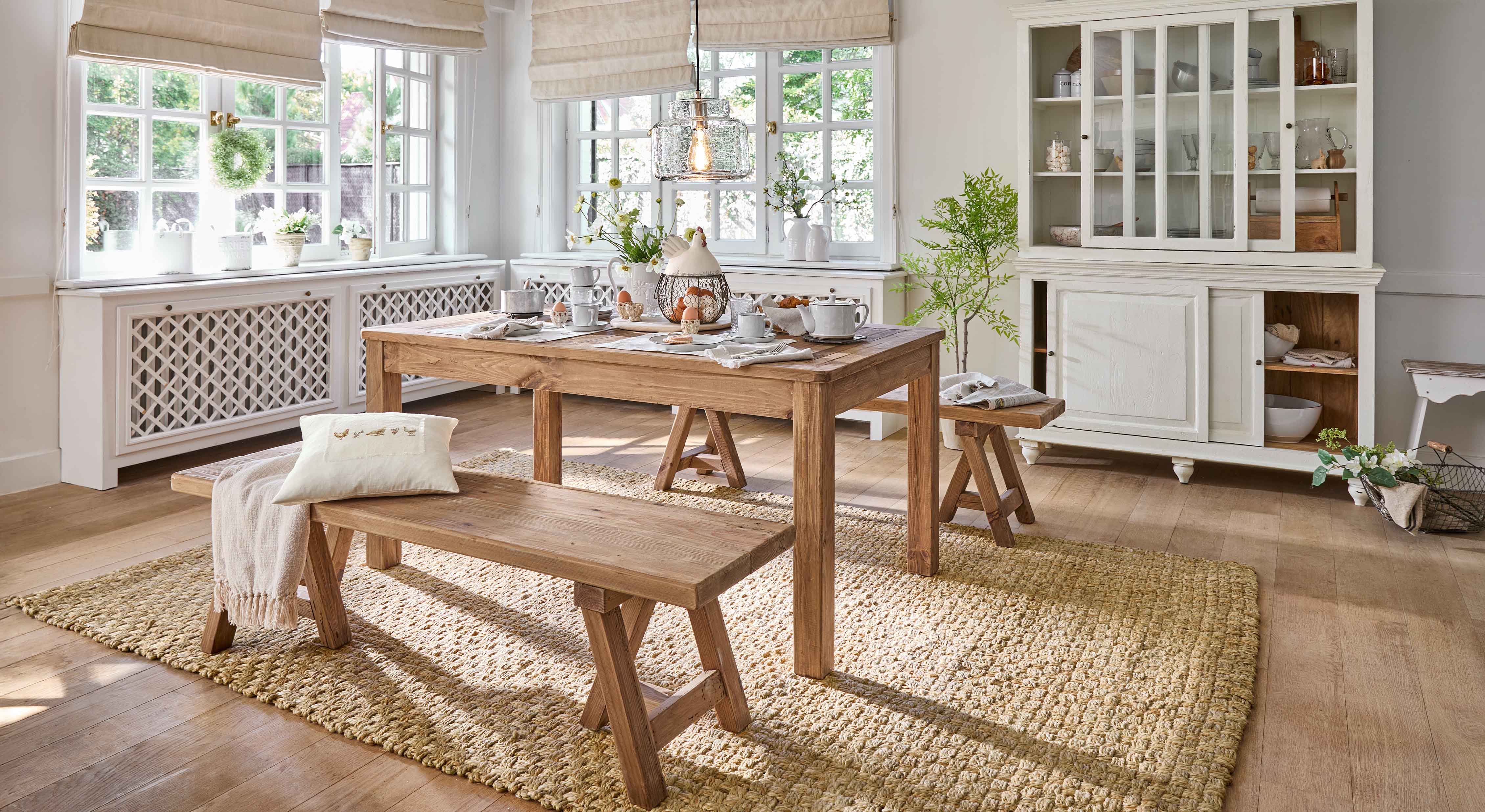 Esszimmer mit Holztisch und Bänken auf Naturteppich, helles Interieur mit weißem Vitrinenschrank