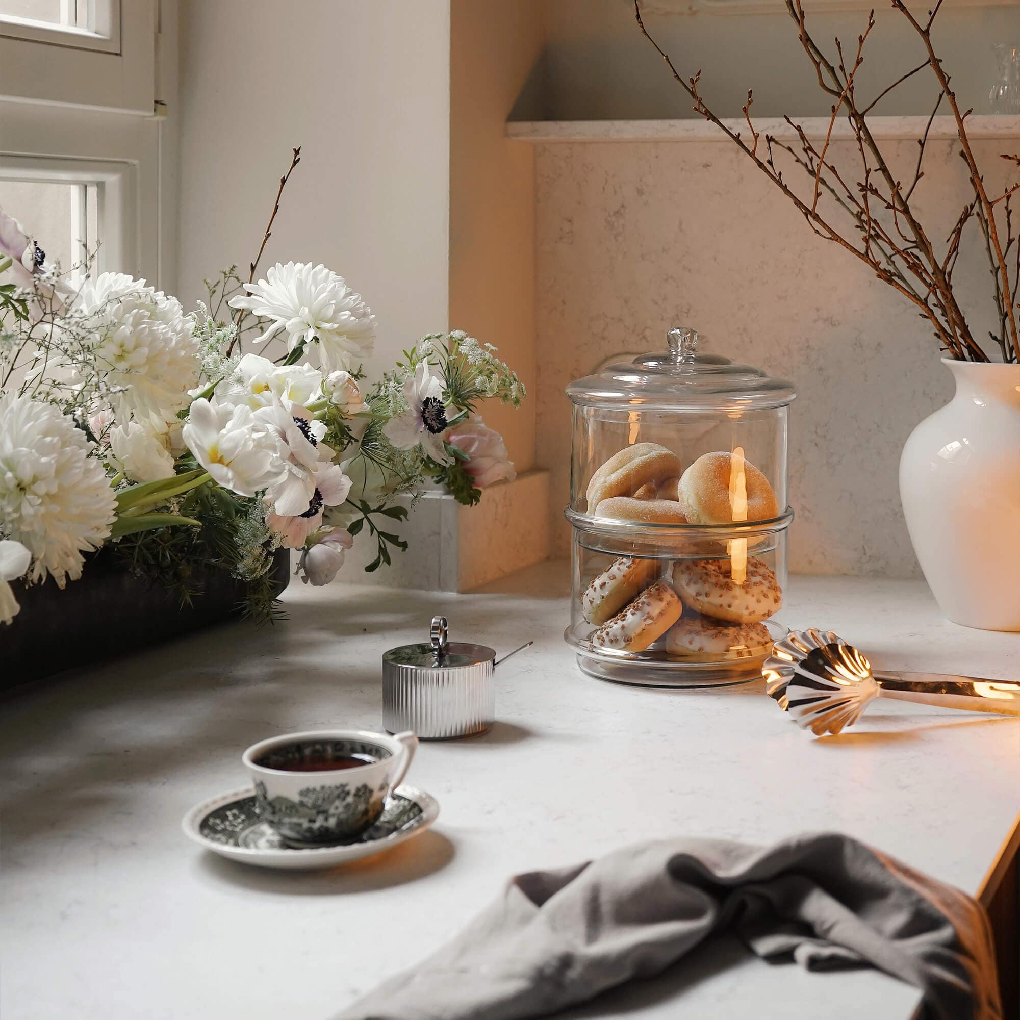Glasbehälter mit zwei Etagen für Gebäck auf hellem Küchenarbeitsplatz mit Blumen und Teetasse