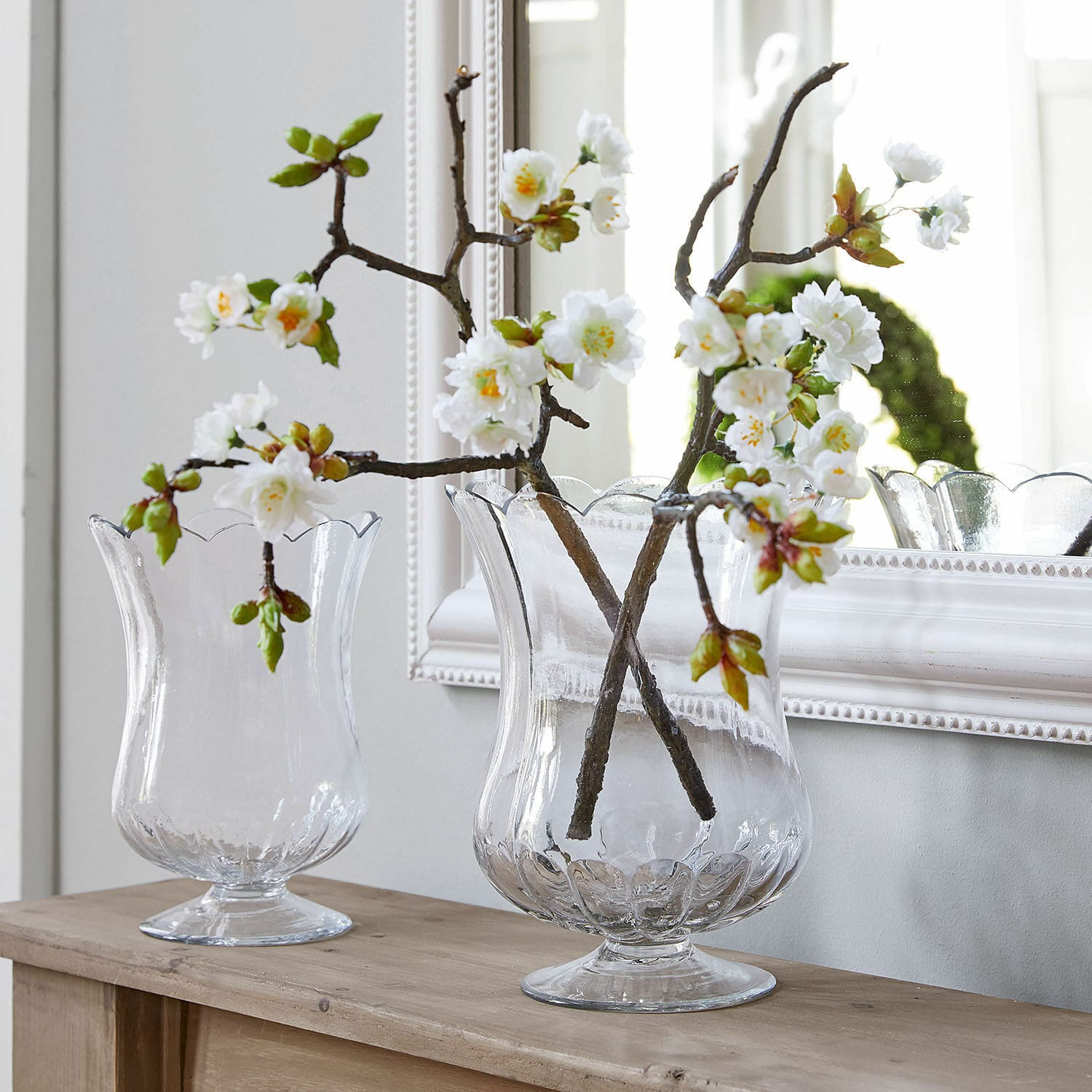 Dekoblume 2er Set Gráda Transparente Glasvasen mit welligem Rand, auf einem Holztisch mit weissen Dekoblumen arrangiert