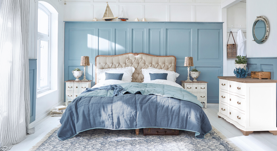 Romantisches Schlafzimmer mit blauer Wand, beige gepolstertem Bett und weissen Holzmöbeln in hellem Ambiente.