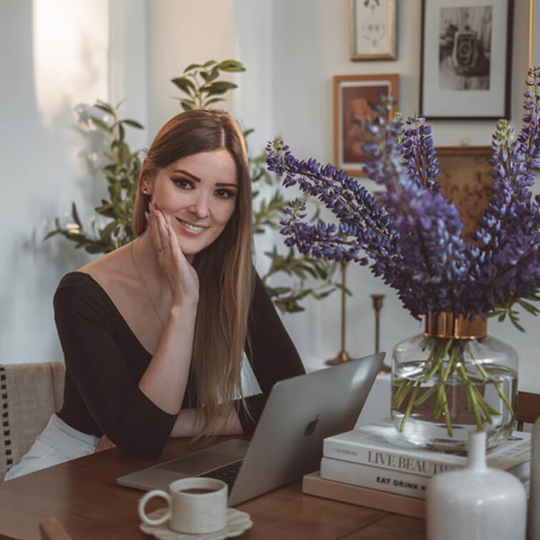 Frau arbeitet am Laptop an Holztisch mit lila Blumen in Glasvase und Kaffeetasse in gemütlicher Umgebung