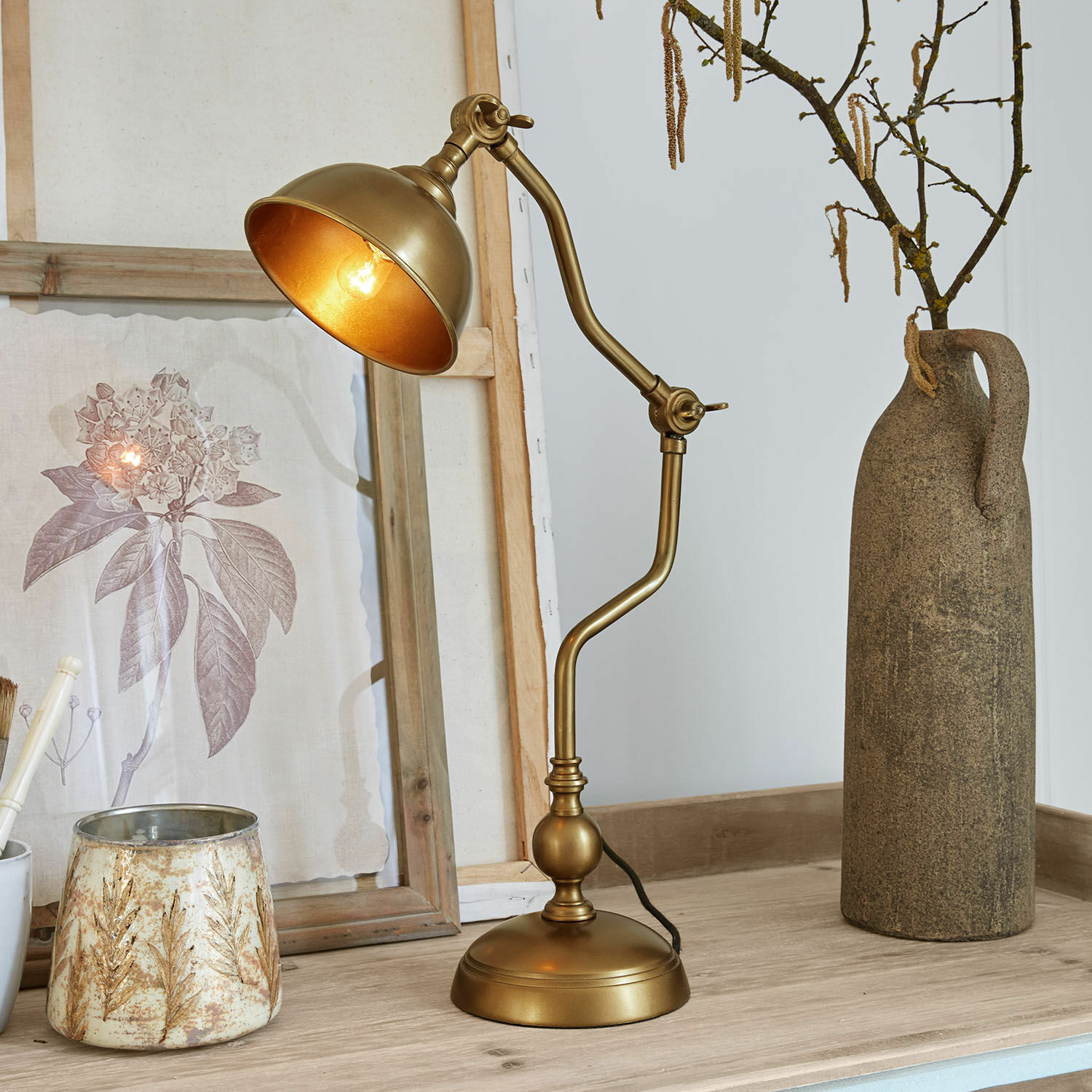 Tischlampe Harrowden Goldfarbene Metall-Tischlampe mit verstellbarem Arm auf Holzplatte mit Dekorationen.
