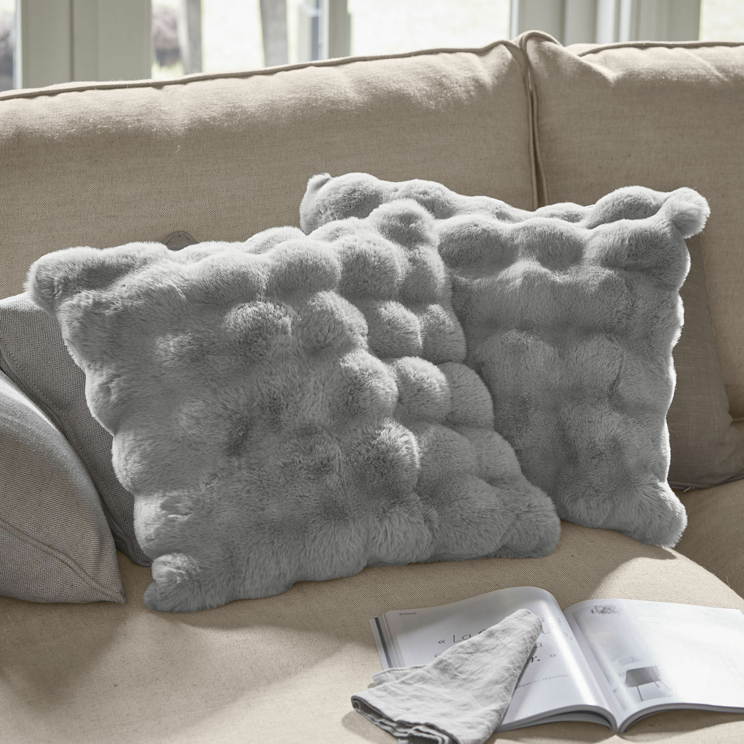 Kissenhülle 2er Set Zalina Weiche, graue Dekokissen aus Fellimitat auf beigem Sofa, ideal für ein gemütliches Wohnzimmer.