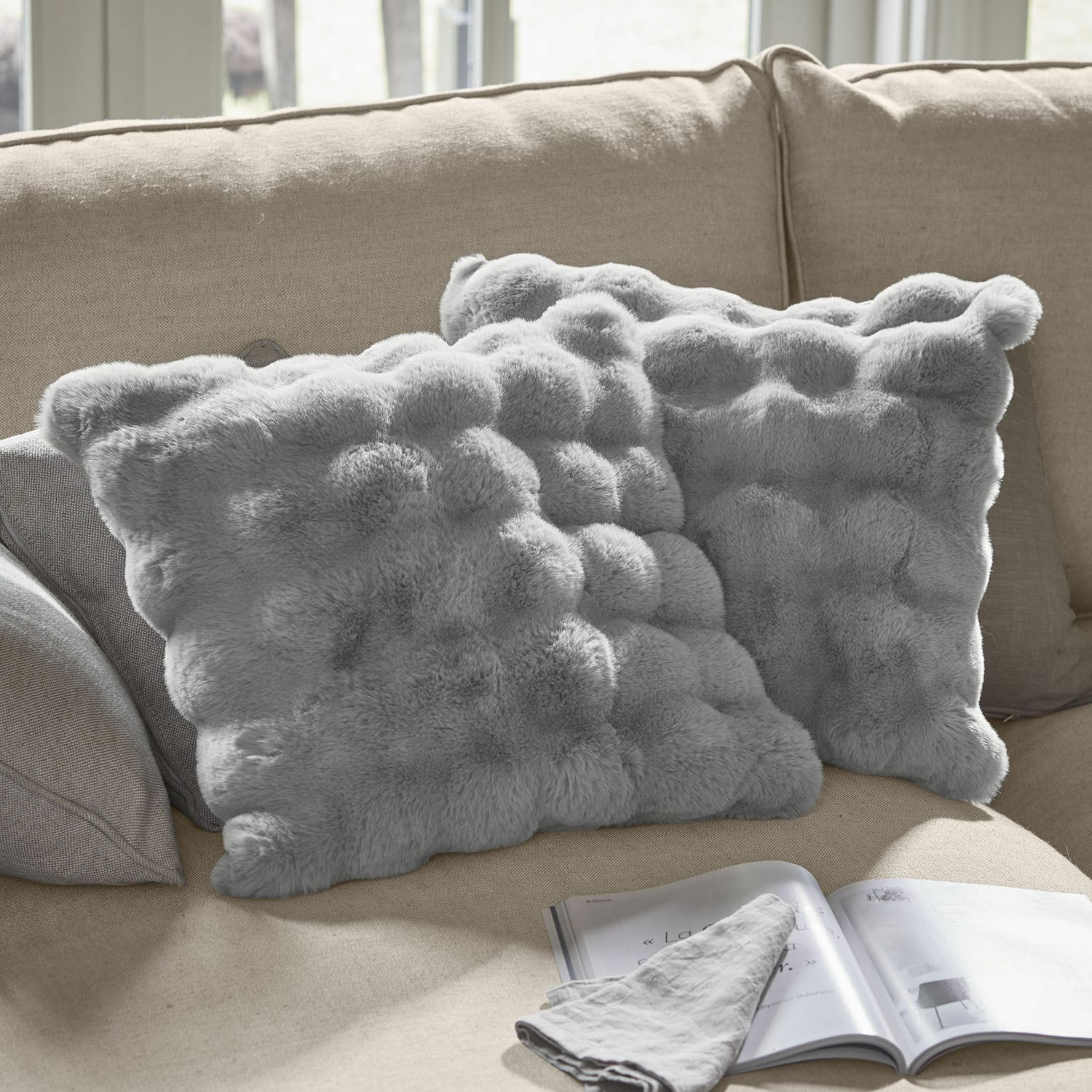 Kissenhülle 2er Set Zalina Weiche, graue Dekokissen aus Fellimitat auf beigem Sofa, ideal für ein gemütliches Wohnzimmer.