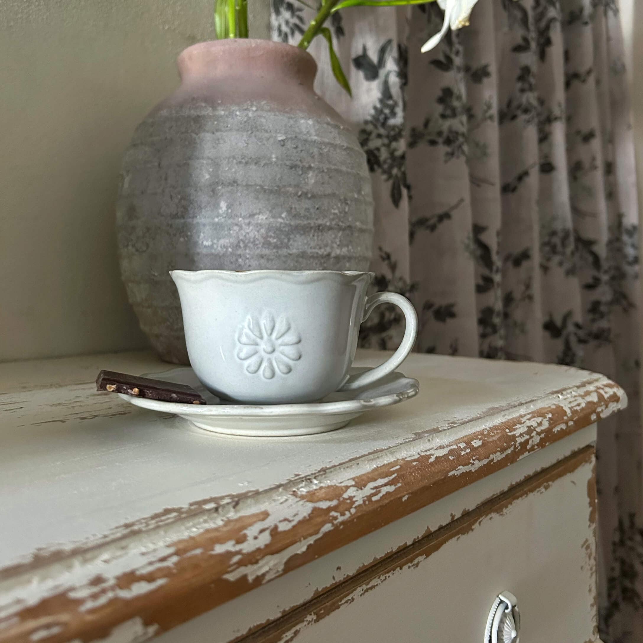Weisse Keramiktasse mit Unterteller und Schokoladenstück auf abgenutztem Holztisch