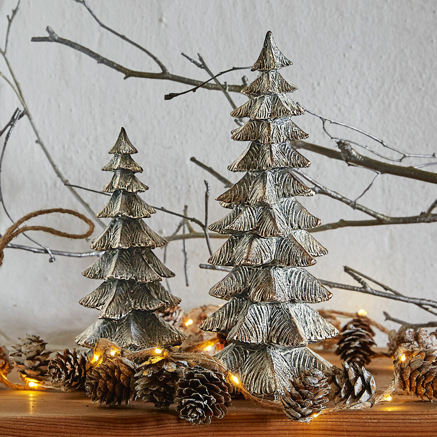 Deko-Baum 2er Set Calavi Dekorative silberne Metall-Weihnachtsbäume auf Holztisch mit Lichterkette und Tannenzapfen.