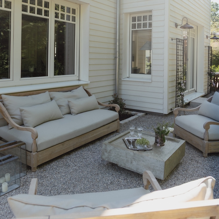 Gartenmöbel-Set aus hellem Holz mit grauen Polstern auf Kiespatio vor weissem Haus
