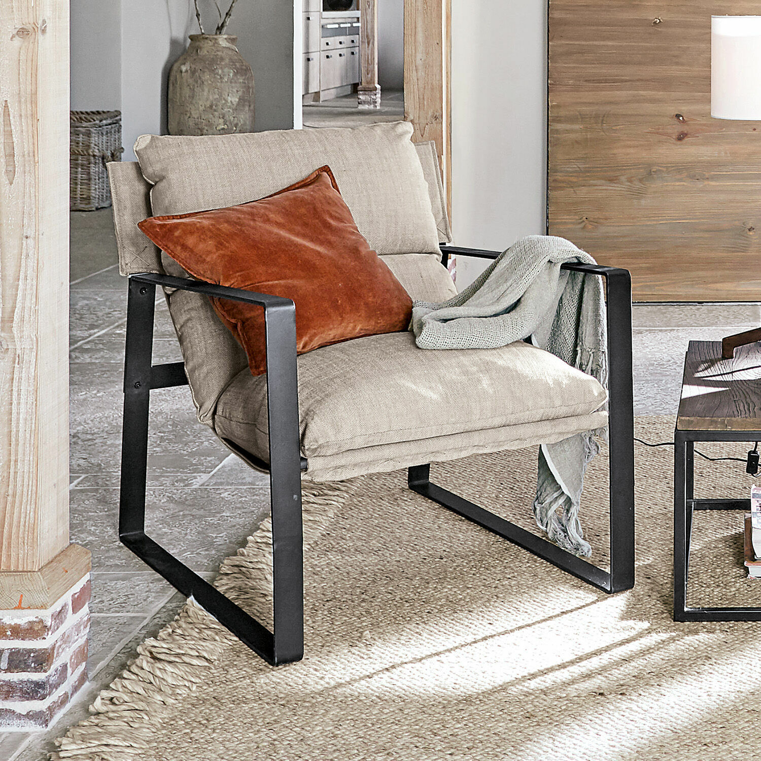 Sessel Seuvard Beiger Polsterstuhl mit schwarzem Metallgestell, orangefarbenem Kissen und grauem Plaid im Wohnzimmer.