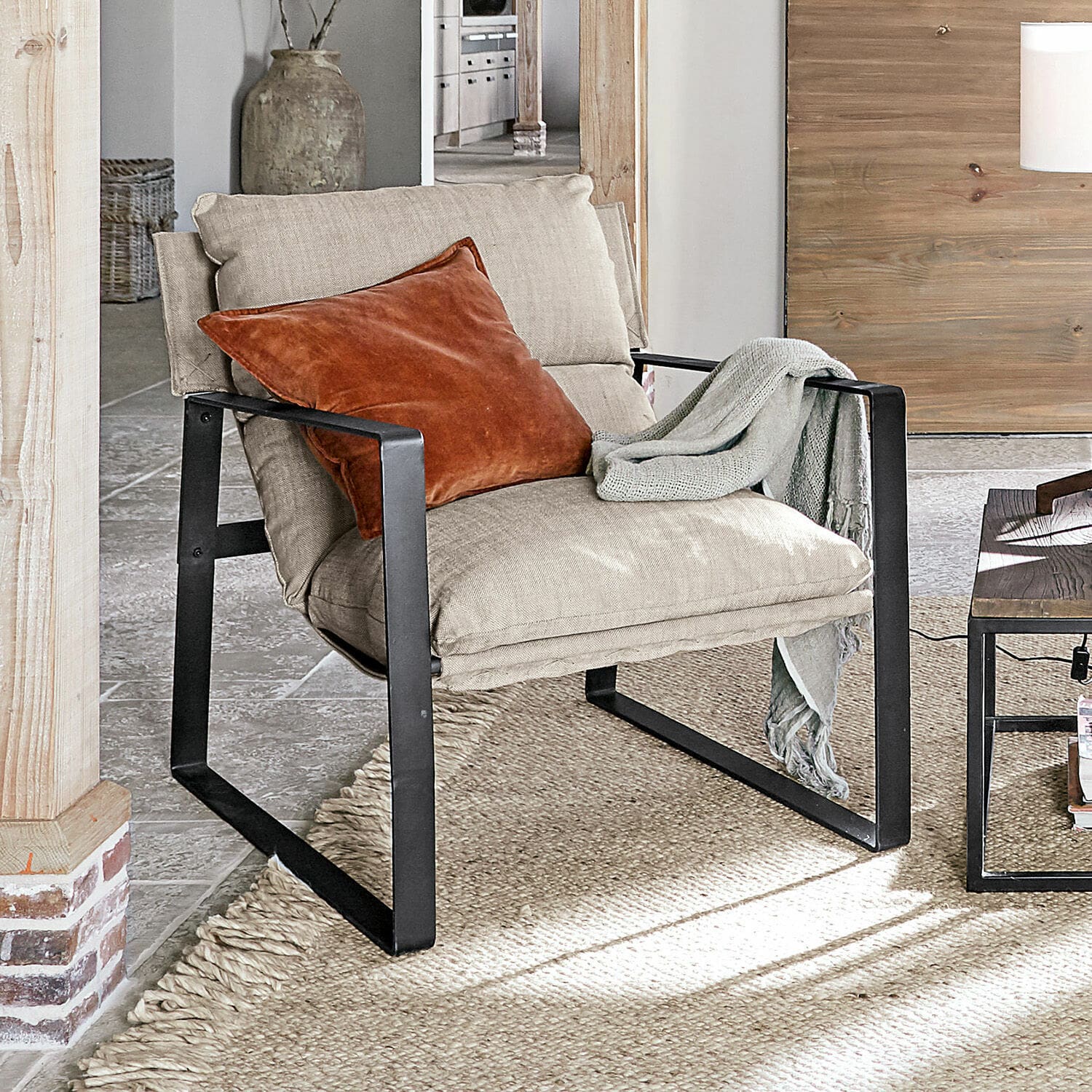 Sessel Seuvard Beiger Polsterstuhl mit schwarzem Metallgestell, orangefarbenem Kissen und grauem Plaid im Wohnzimmer.