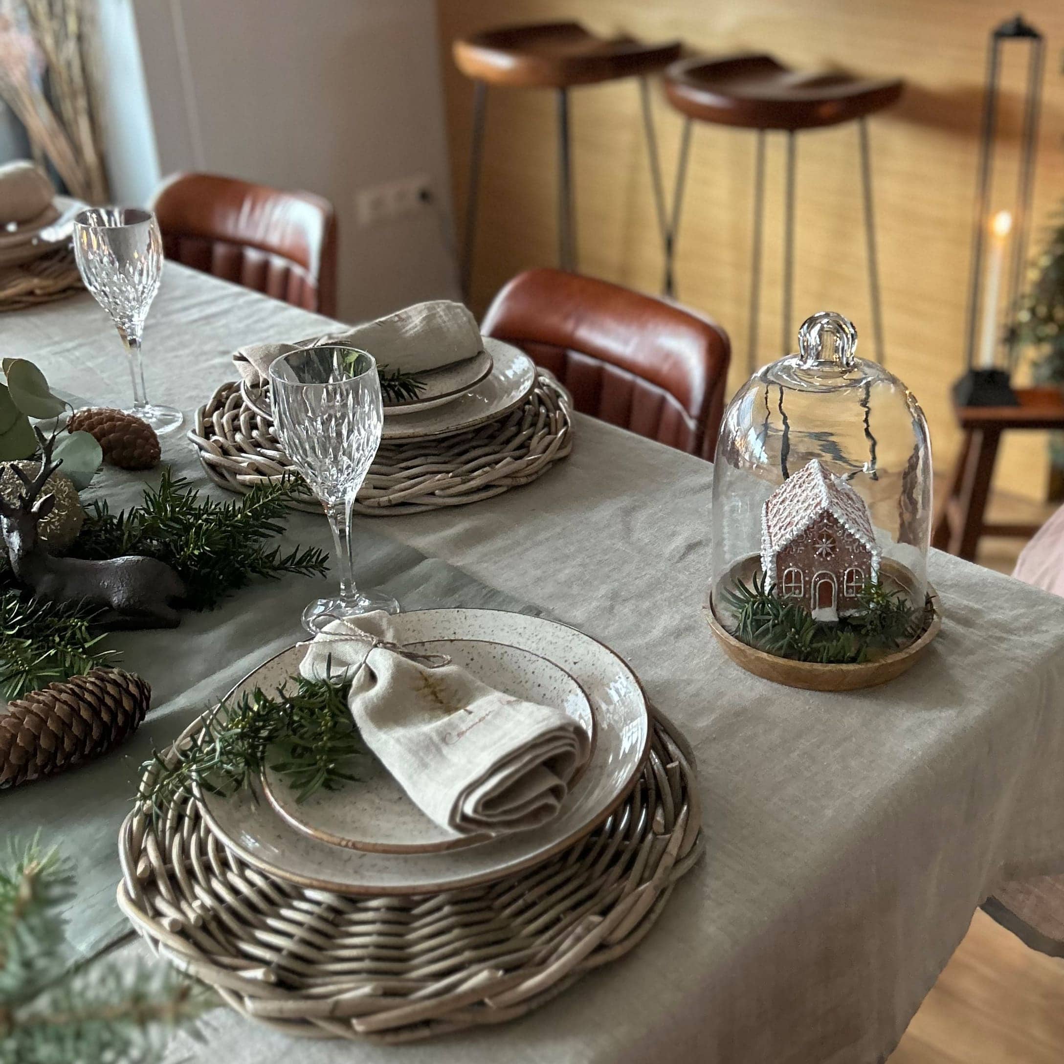 Eleganter gedeckter Tisch mit geflochtenen Platzsets, Keramiktellern und Weihnachtsdekoration mit Häuschen unter Glaskuppel