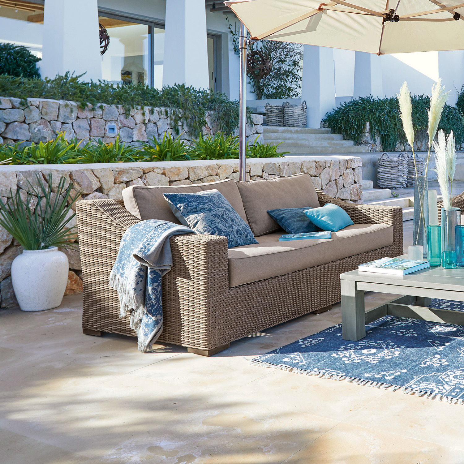 Beiges Rattan-Gartensofa mit blauen Kissen und Sonnenschirm auf sonniger Terrasse