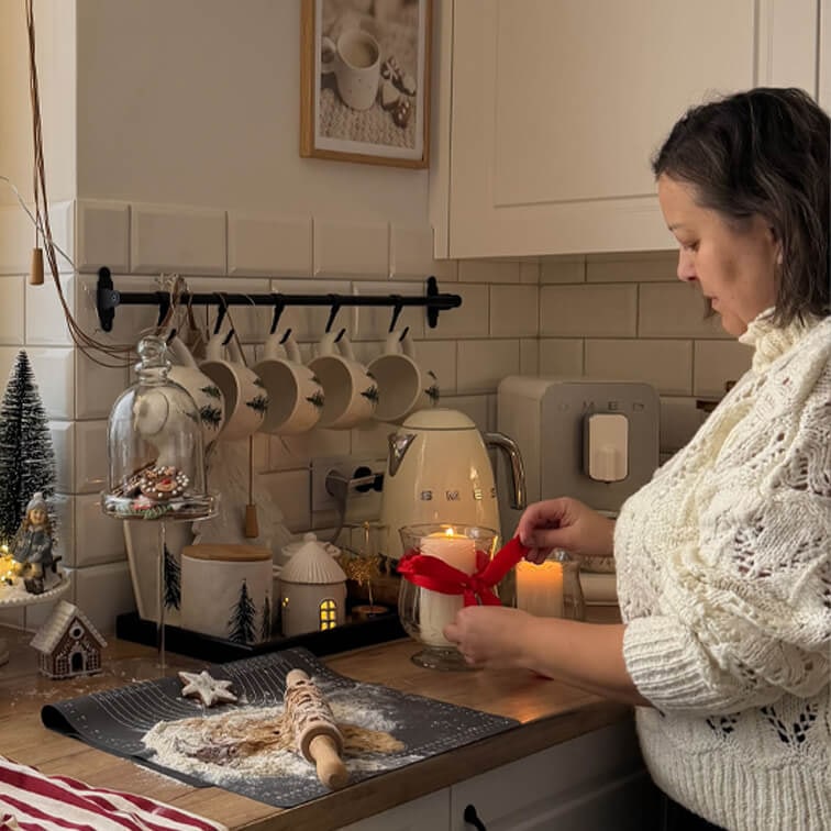 Frau im weissen Pullover bindet rote Schleife um Kerze in gemütlicher Küche mit Backutensilien