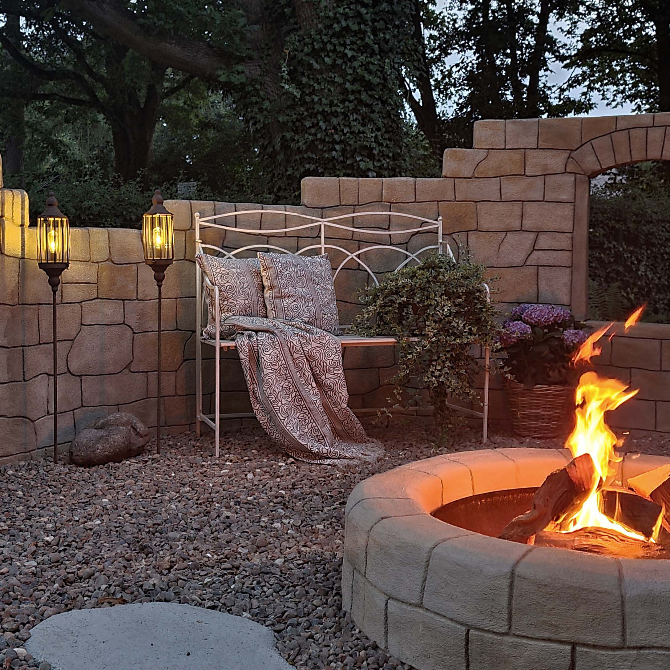 Garten mit steinernem Feuerplatz, weisser Metallbank und Stehlampen auf Kies für den Aussenbereich