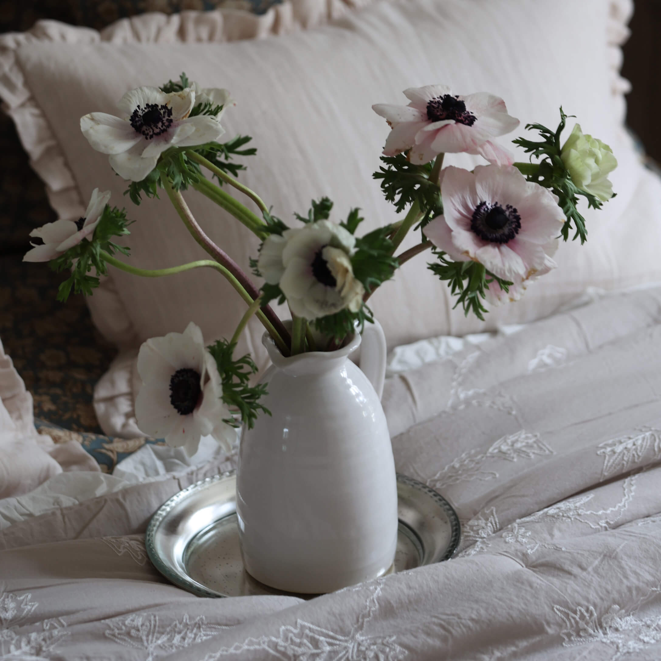 Weisser Keramikkrug mit zarten weissen Anemonen auf bestickter beiger Bettwäsche