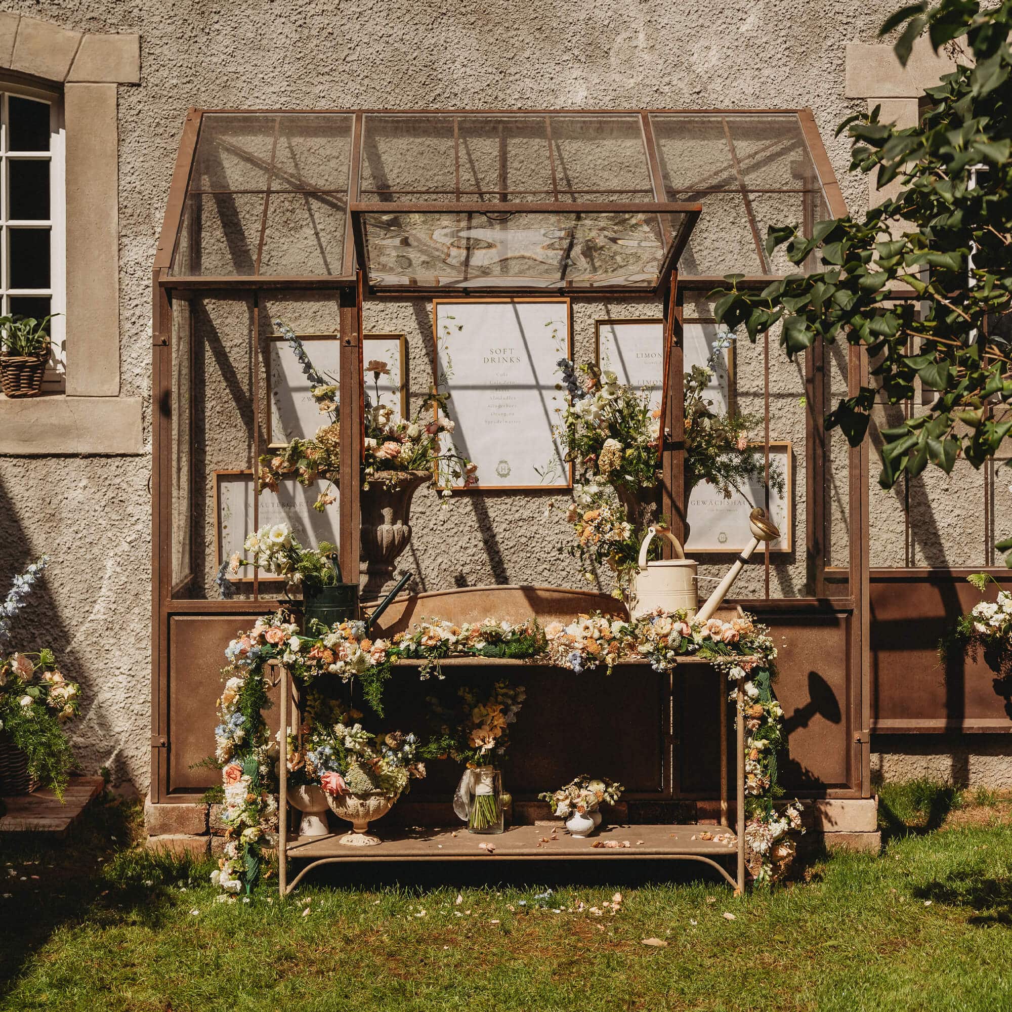 Metall-Gartentisch mit Blumenarrangements und Giesskannen vor einer Glashauswand
