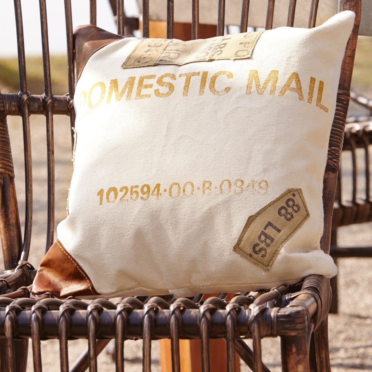 Kissen Farway Beiges Kissen mit Aufdruck Domestic Mail und braunen Lederecken auf einem Rattanstuhl