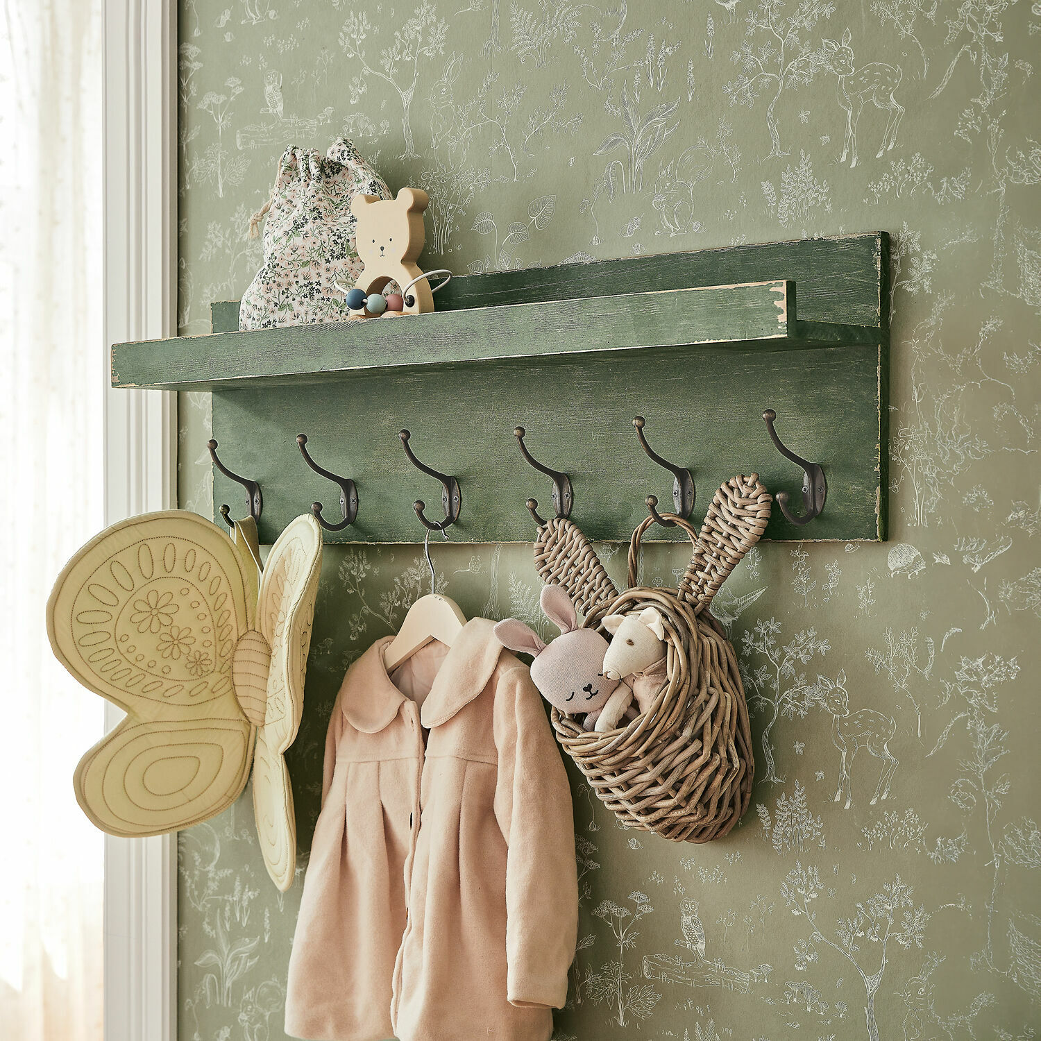 Wandregal Hoboken Grüne Holz-Wandgarderobe mit Haken, Kinderjacke und Korb mit Plüschtieren vor Tapete mit Tiermuster