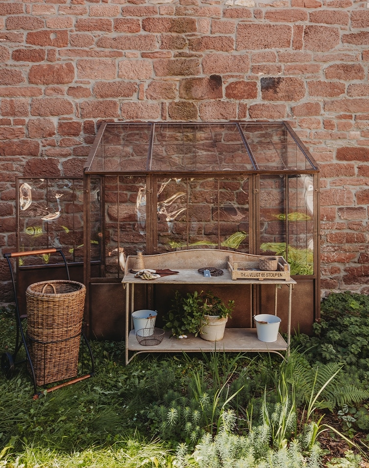 Garten-Gewächshaus aus Glas mit Metallrahmen und Holztisch vor roter Ziegelmauer