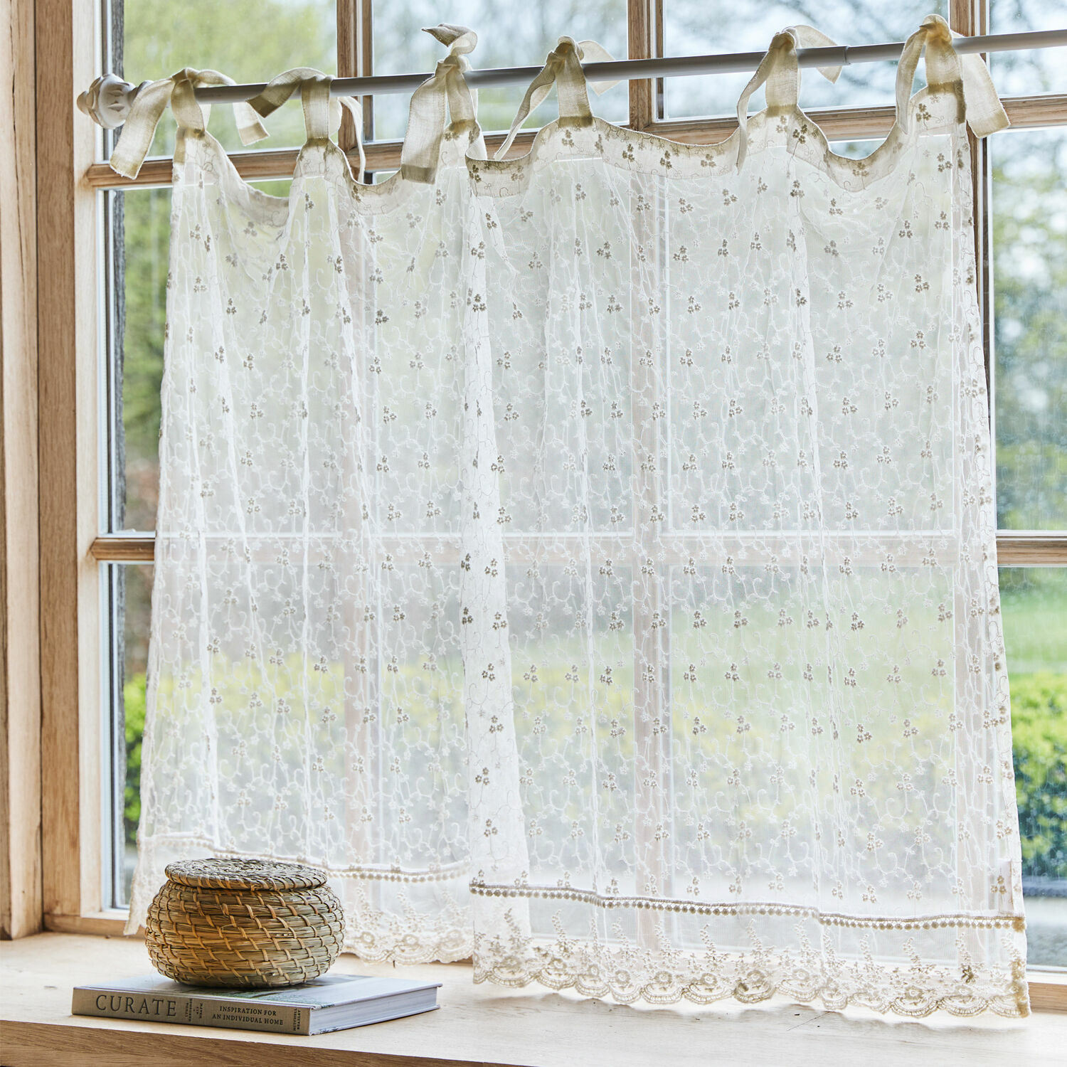 Scheibengardine Auradé Transparenter, bestickter cremefarbener Vorhang mit Schleifen an einem Fenster mit Gartenblick.