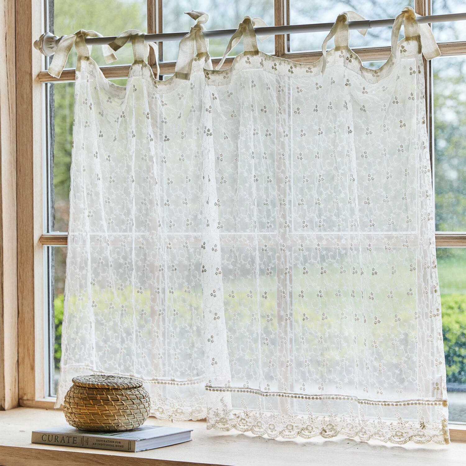 Scheibengardine Auradé Transparenter, bestickter cremefarbener Vorhang mit Schleifen an einem Fenster mit Gartenblick.