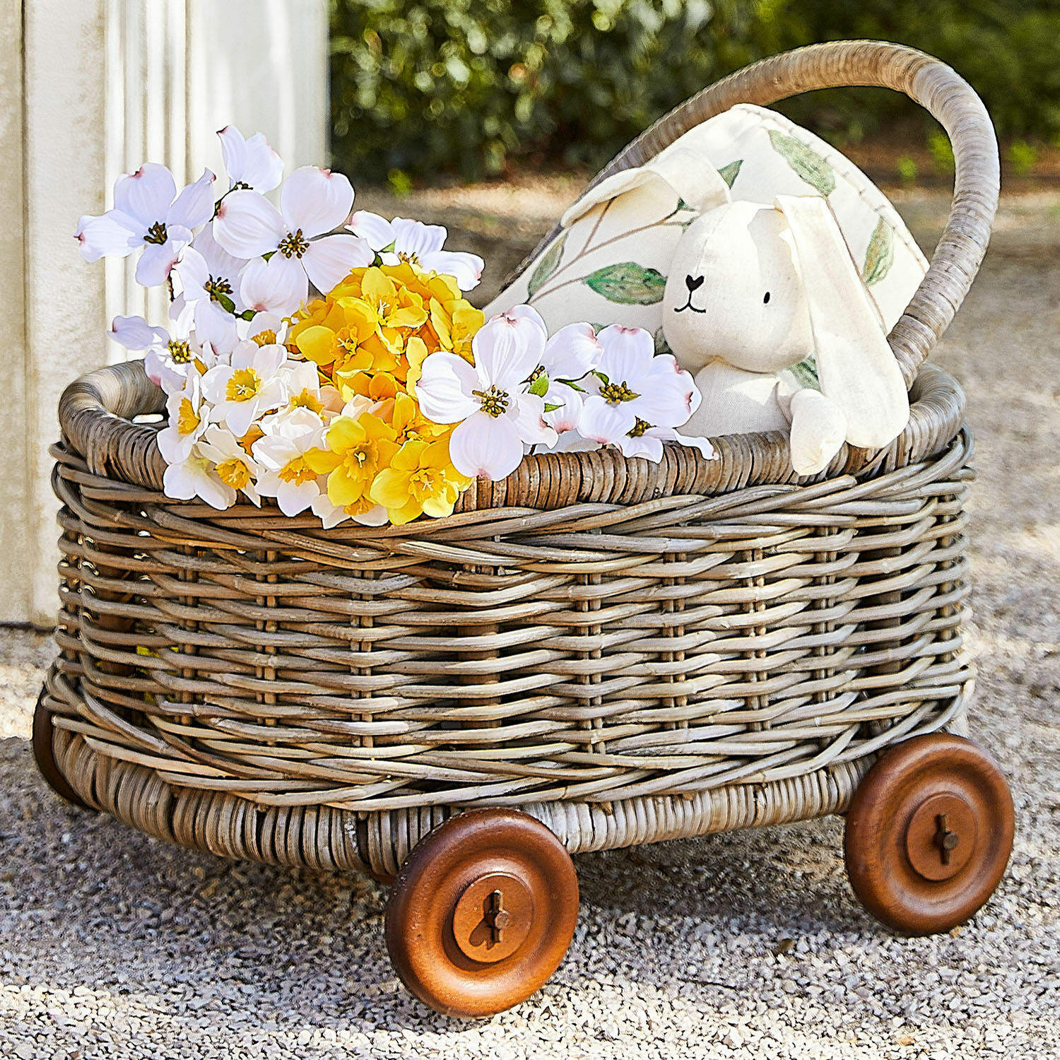 Rollwagen Narivena Wagen aus Rattan mit Holzrädern, gefüllt mit gelben und weissen Blumen und einem Plüschhasen