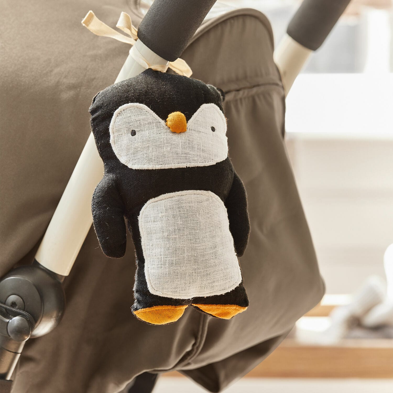 Kinderwagenanhänger Jocienne Schwarzer Plüsch-Pinguin mit weissem Bauch und orangen Füssen am Kinderwagen befestigt
