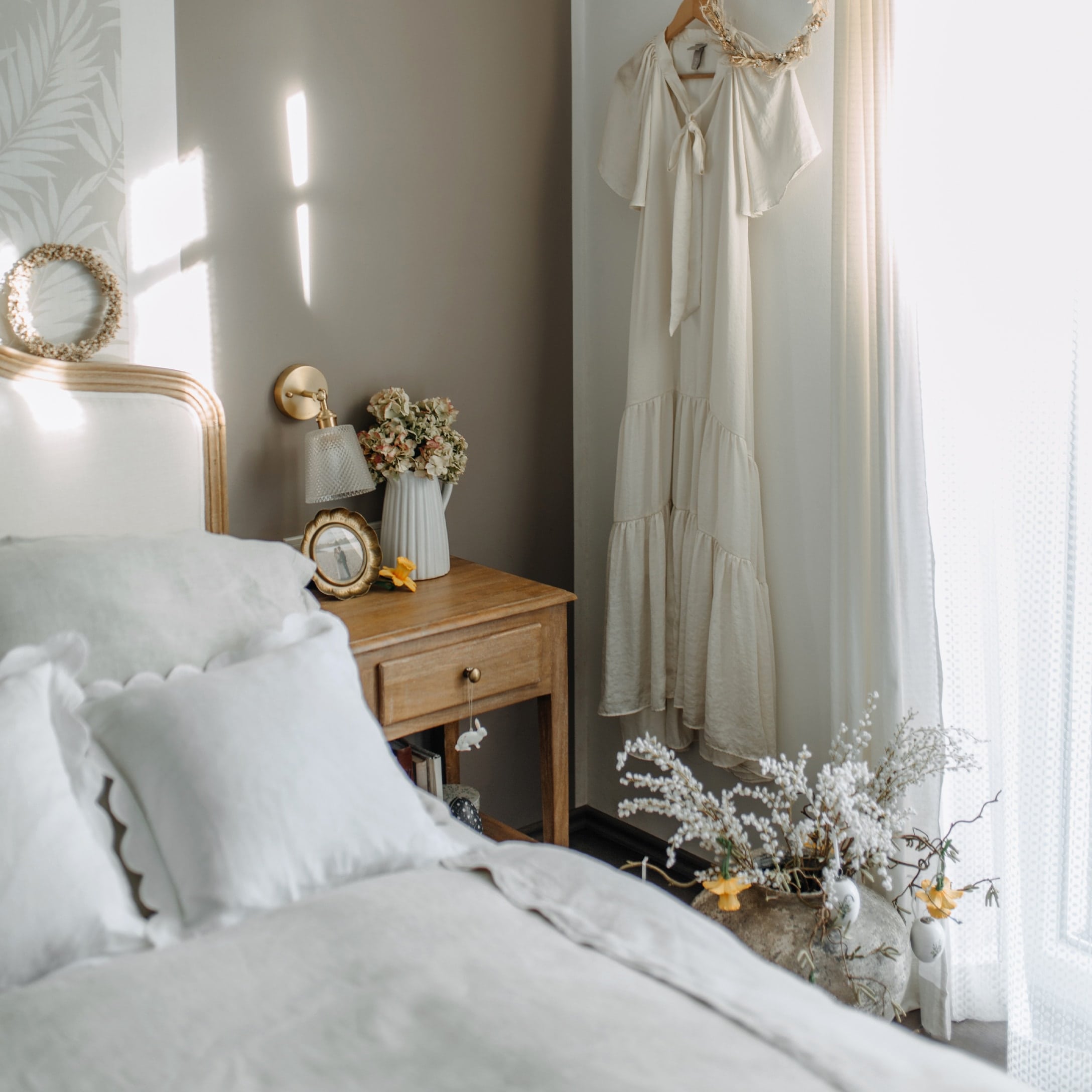 Gemütliches Schlafzimmer mit Holznachttisch, weisser Bettwäsche und cremefarbenem Kleid an der Wand