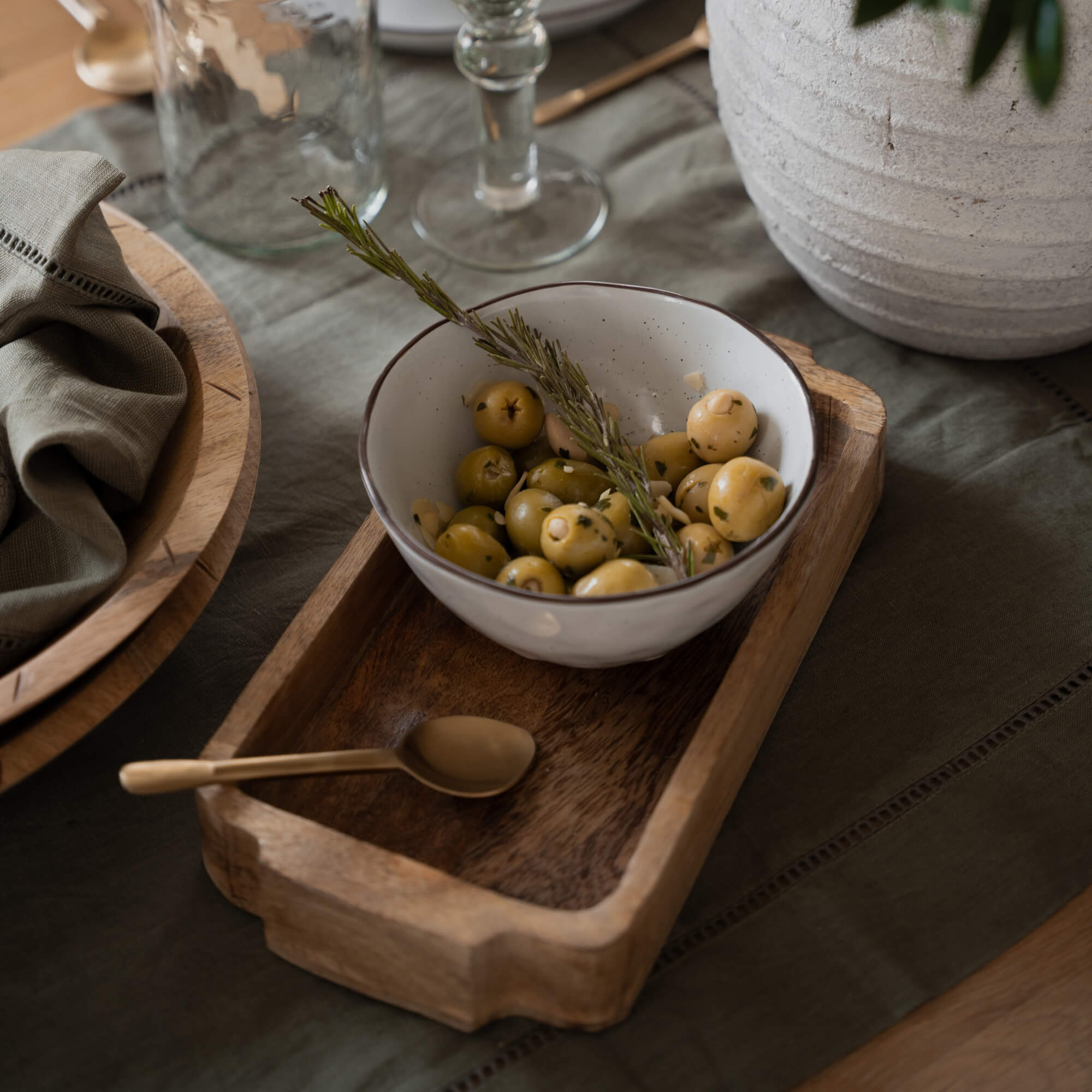 Weisse Keramikschale mit grünen Oliven und Rosmarinzweig auf Holztablett auf dem Tisch