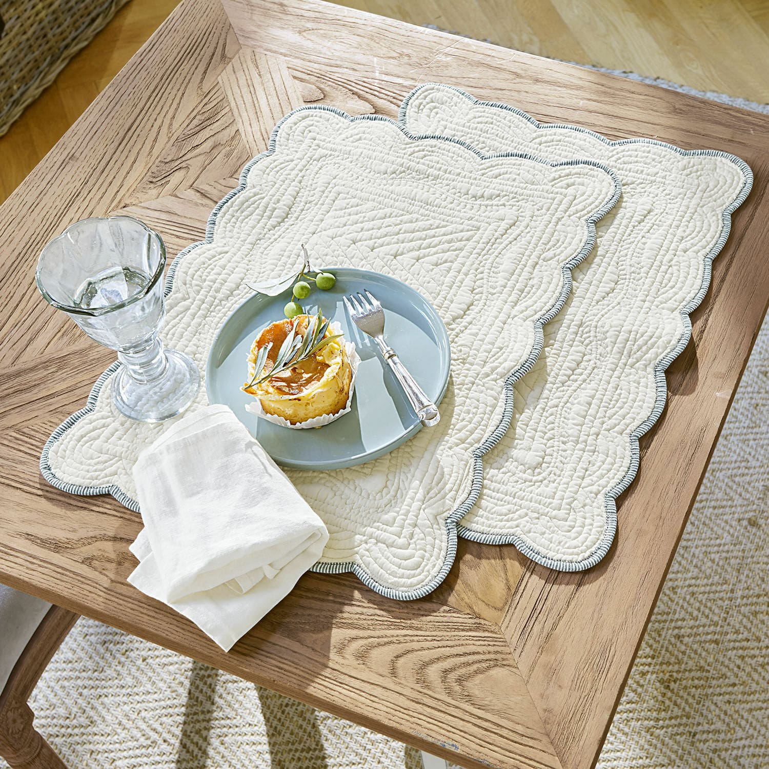 Platzset 2er Set Fontienne Beige gesteppte Baumwoll-Tischsets mit blauem Rand auf Holztisch mit Dessert und Glas