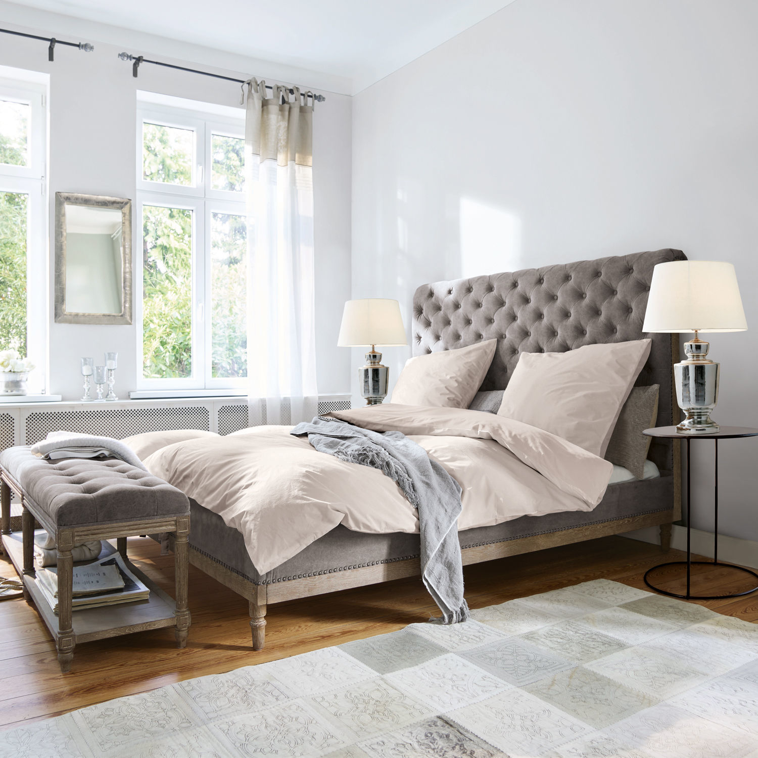 Bett Pozières Grau gepolstertes Bett mit gesteppter Rückenlehne und beiger Bettwäsche in hellem Schlafzimmer mit Holzboden