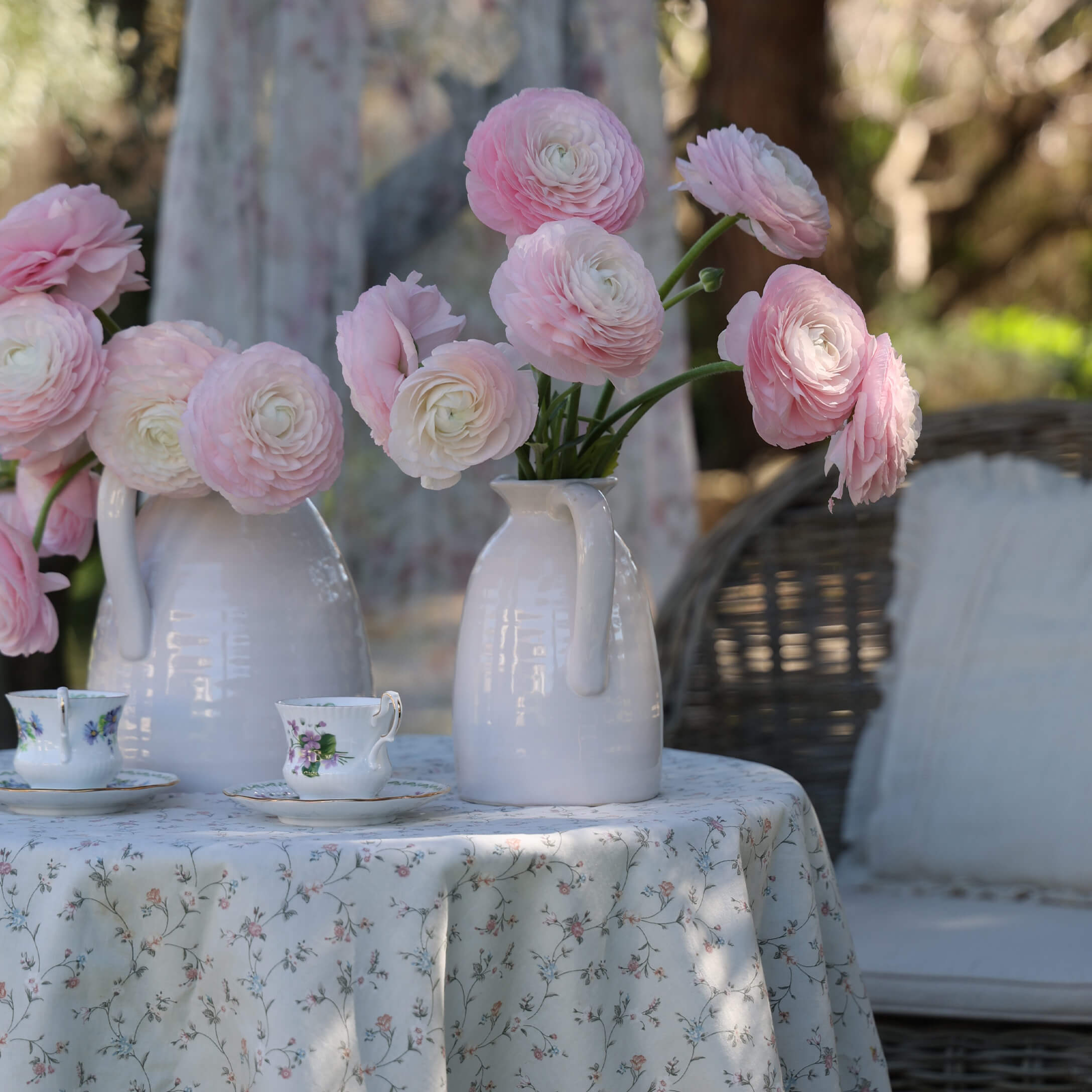 Weiße Keramikkrüge mit rosa Blumen auf einem Tisch mit geblümtem Tischtuch und dekorativen Teetassen