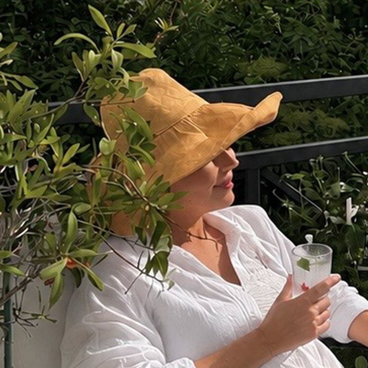 Frau in weisser Bluse und gelbem Strohhut entspannt auf Terrasse mit Getränk in der Hand