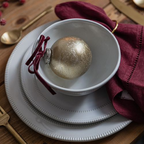 Weisses Keramik-Teller-Set mit bordeauxroter Stoffserviette und goldener Kugel auf Holztisch