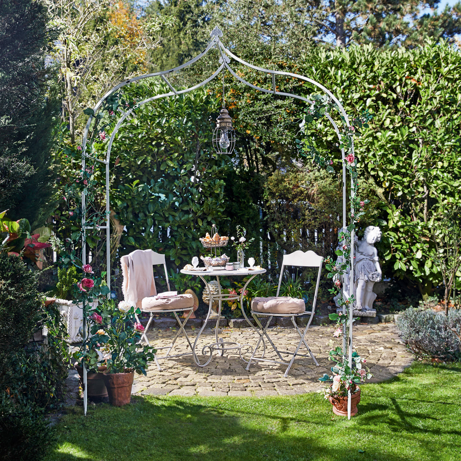 Rosenbogen Dimarus Weisser Metall-Gartenbogen mit rosa Blumen und Hängelampe, davor Sitzgruppe auf Steinpflasterung