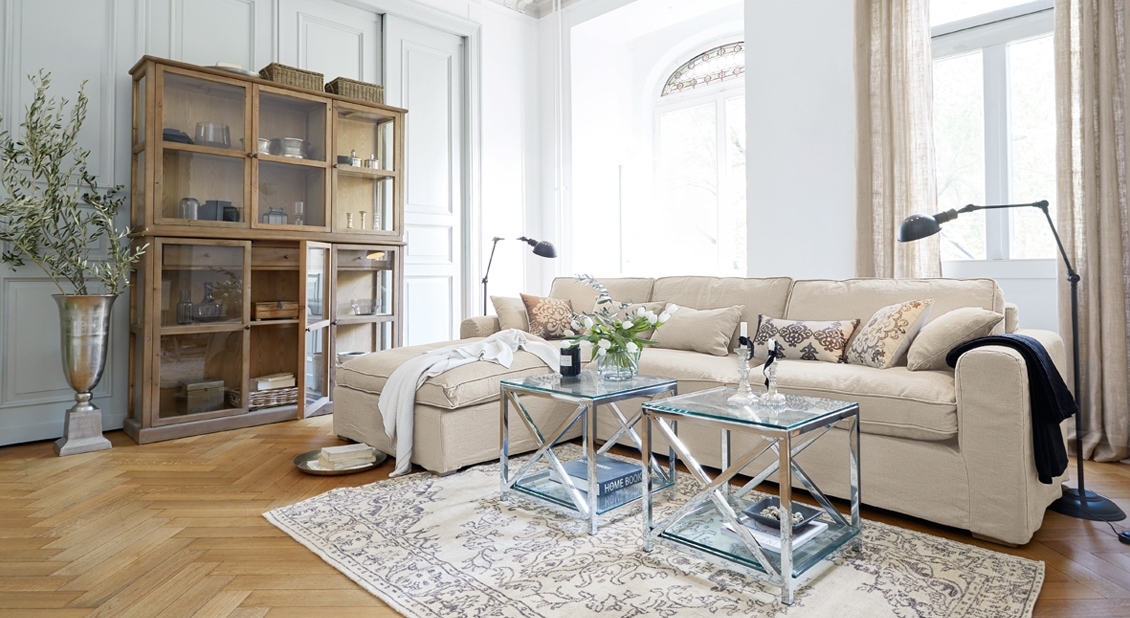Gemütliches Wohnzimmer mit beigem Sofa, Glastischen und Holzvitrine auf hellem Parkettboden.