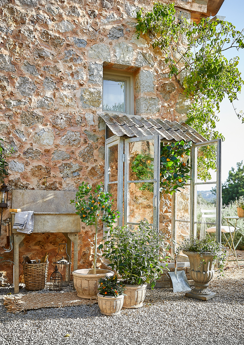 Garten mit Steinmauer, Metallgewächshaus und Pflanzgefässen auf Kiesboden
