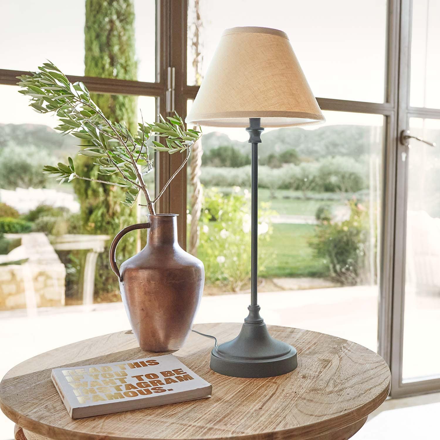 Kupferfarbener Metallkrug mit Zweig und Tischlampe mit beigem Lampenschirm auf Holztisch