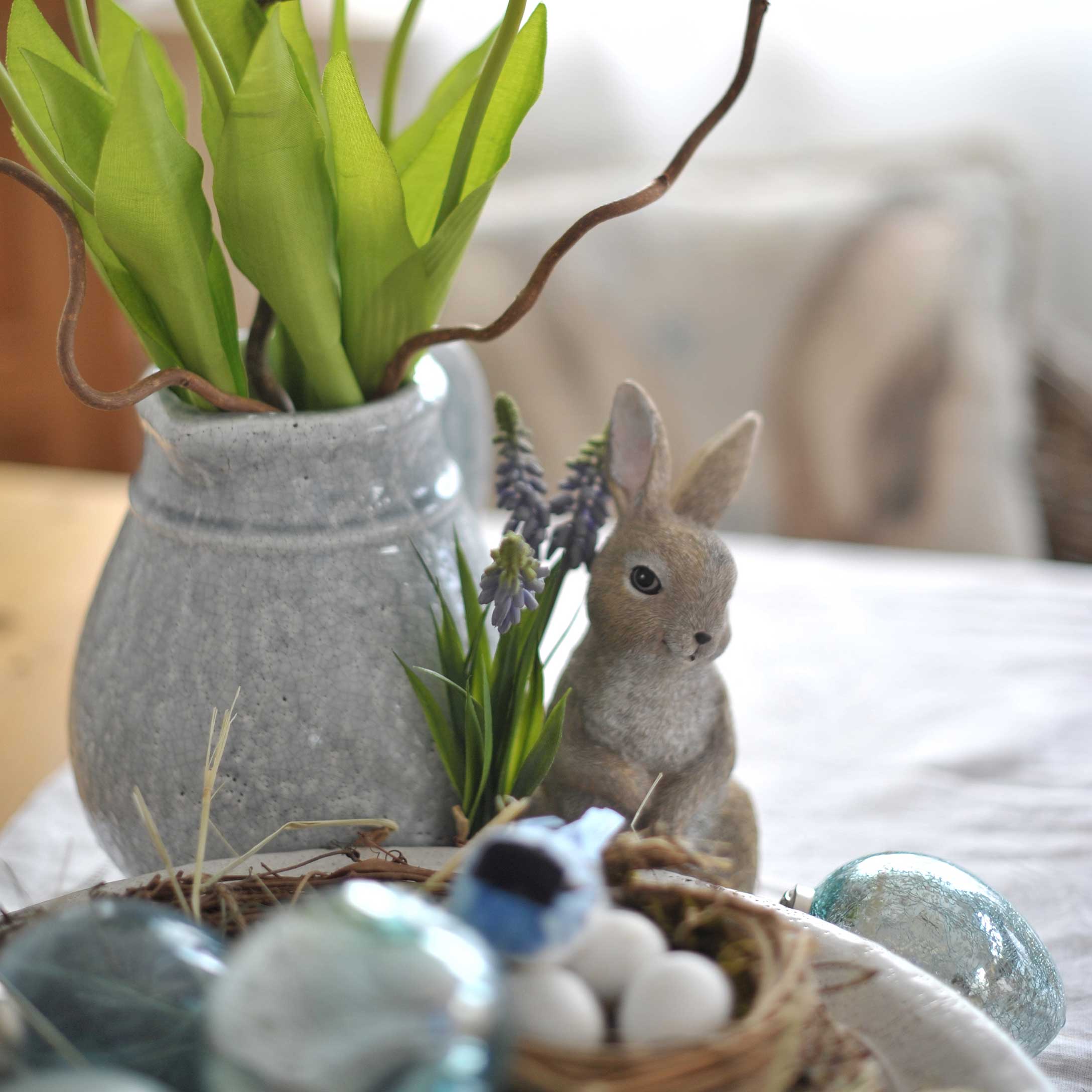 Graue Keramikvase mit grünen Blättern und Hasenfigur als Osterdekoration auf dem Tisch