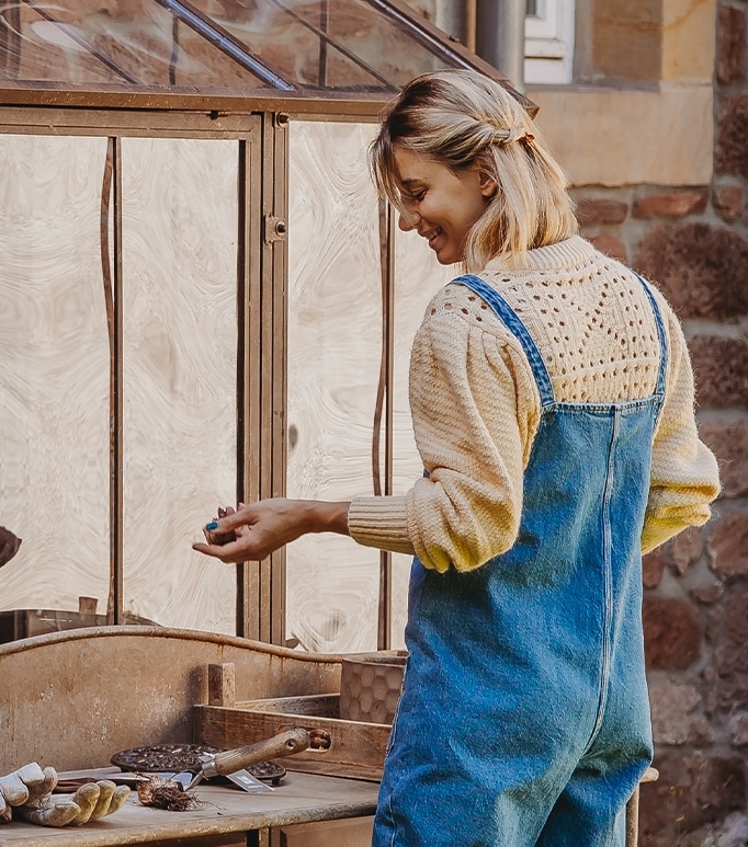 Frau in blauer Latzhose und cremefarbenem Pullover arbeitet an einem Holztisch im Garten.
