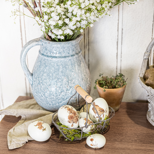Blauer Keramikkrug mit weissen Blumen und dekorativen Eiern mit Tiermotiven auf Holztisch