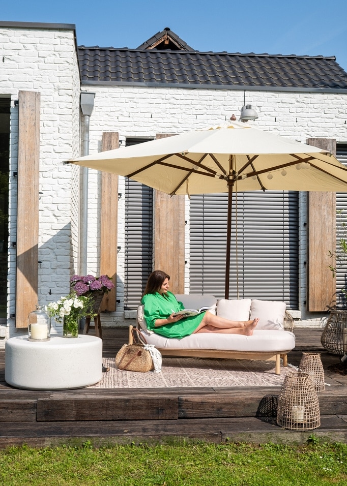 Frau im grünen Kleid entspannt auf hellem Sofa unter cremefarbenem Sonnenschirm auf Holzterrasse.