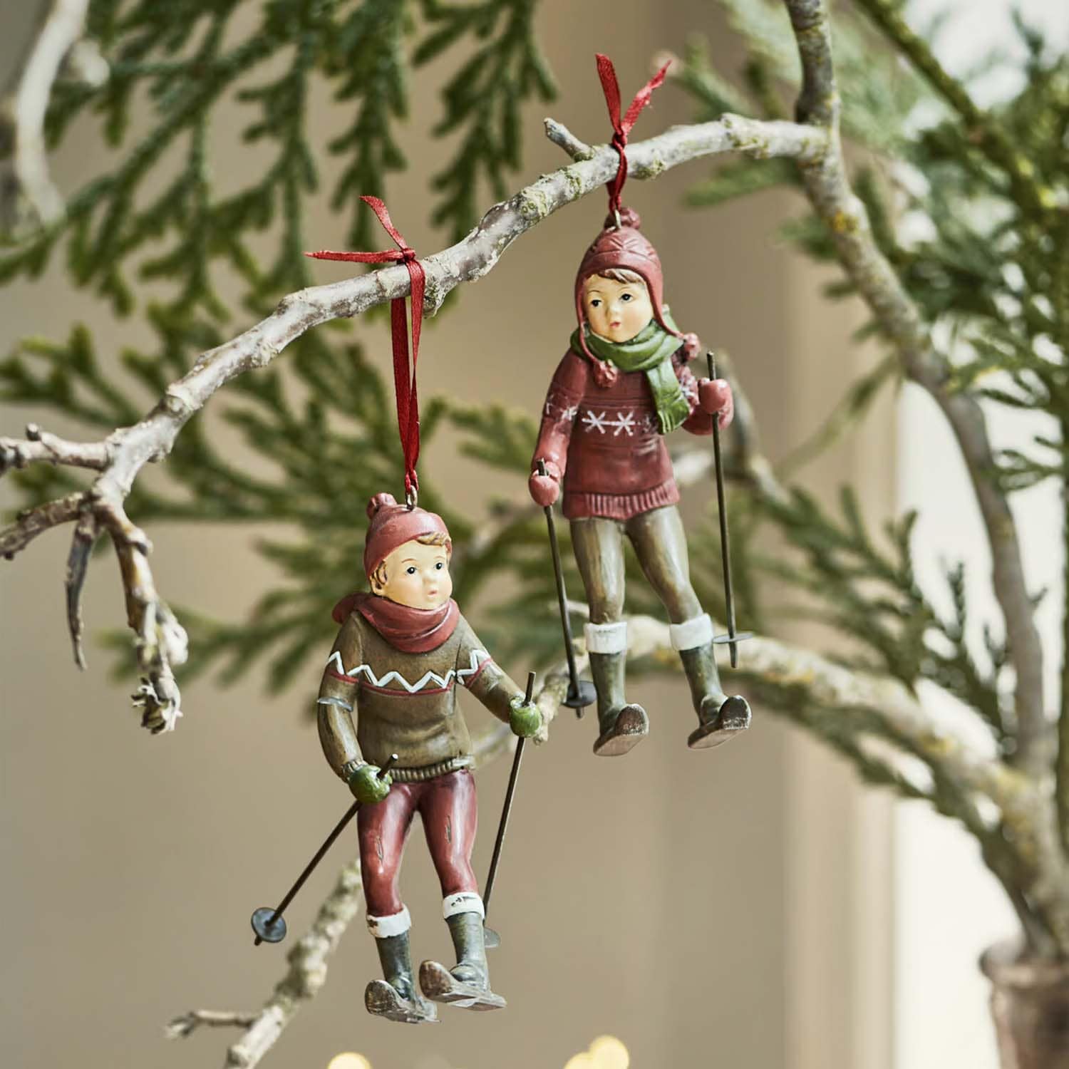 Zwei hängende Figuren von Kindern beim Skifahren in roten und grünen Kleidern an einem Ast