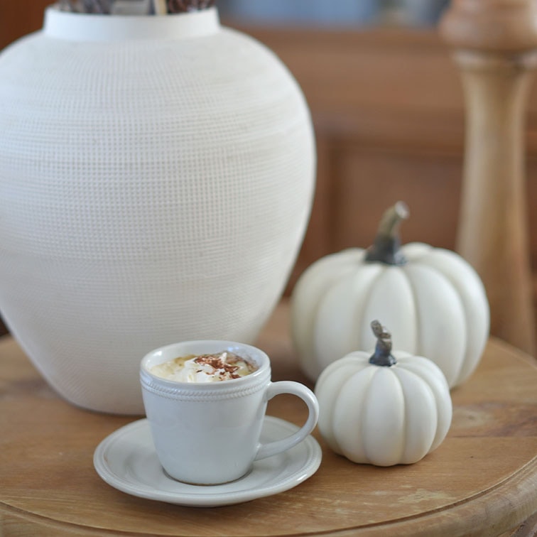 Weiße Keramiktasse mit Kaffee und Sahne auf Holztisch neben weißen Dekokürbissen und Vase