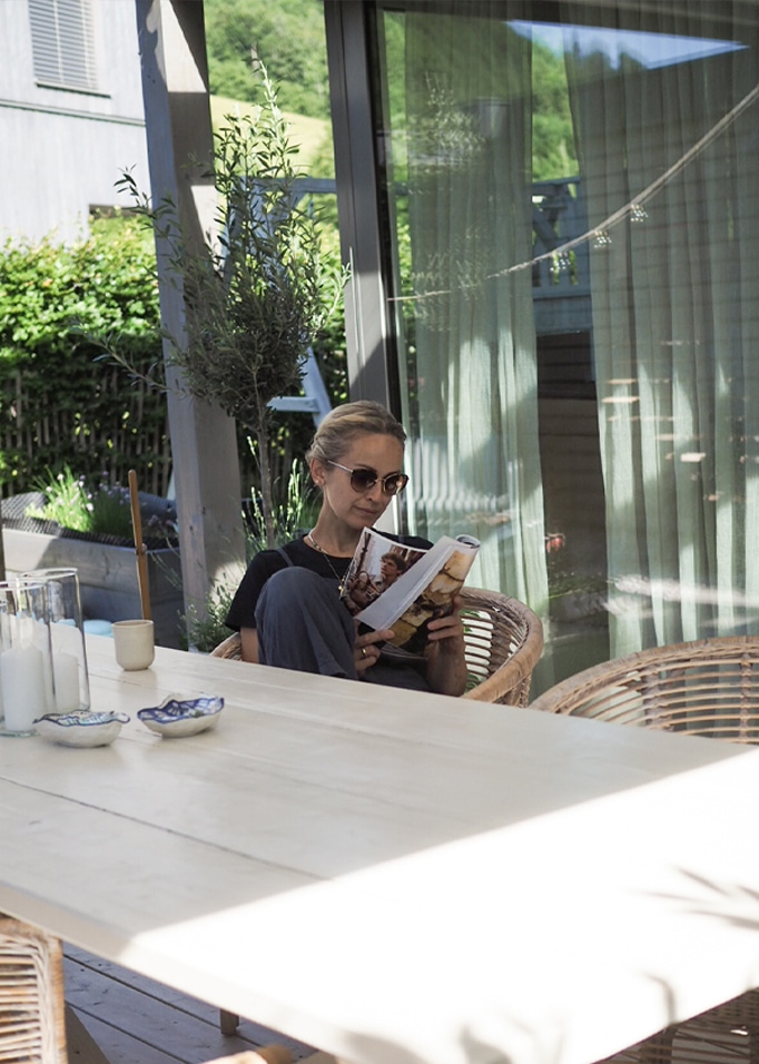 Frau liest Buch auf Terrasse mit hellem Holztisch und Rattanstühlen im Sonnenlicht.