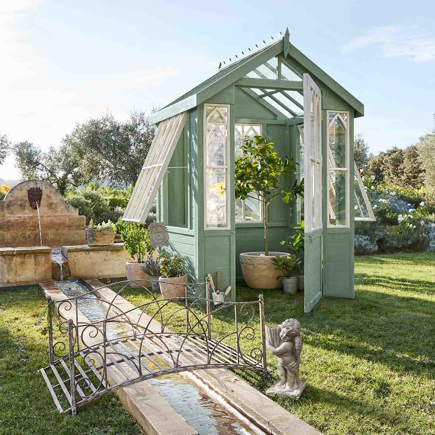 Grünes Holz-Gewächshaus mit weissen Fenstern im Garten mit Pflanzen und Brunnen