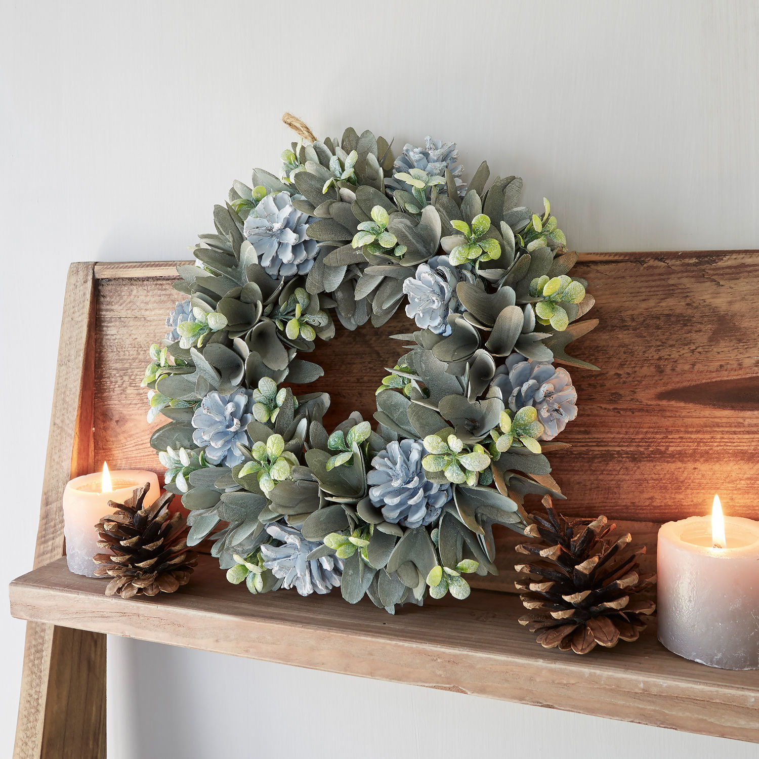 Kranz Deinze Dekorativer Kranz aus grauen Blättern und blauen Tannenzapfen auf Holzregal mit Kerzen und Kiefernzapfen