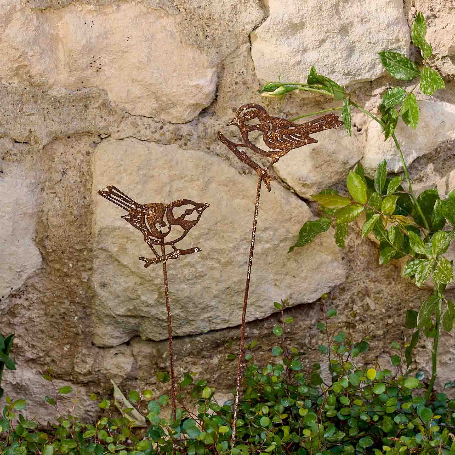 Dekorative Metallvögel mit Rostoptik auf Stäben im Garten vor einer Steinmauer