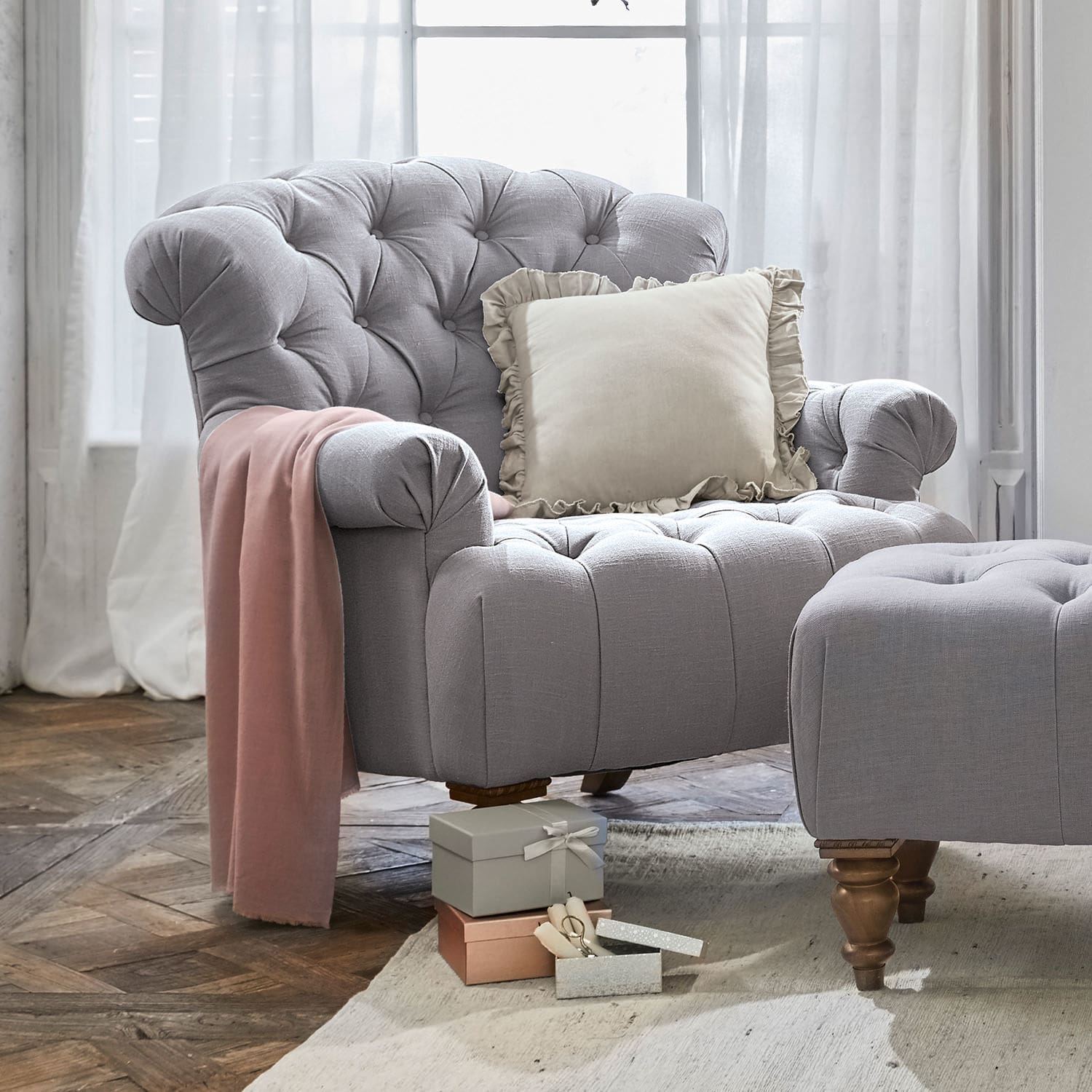 Sessel Springfield Grau gepolsterter Sessel mit Hocker, cremefarbenem Kissen und rosa Decke auf Holzboden im Wohnzimmer